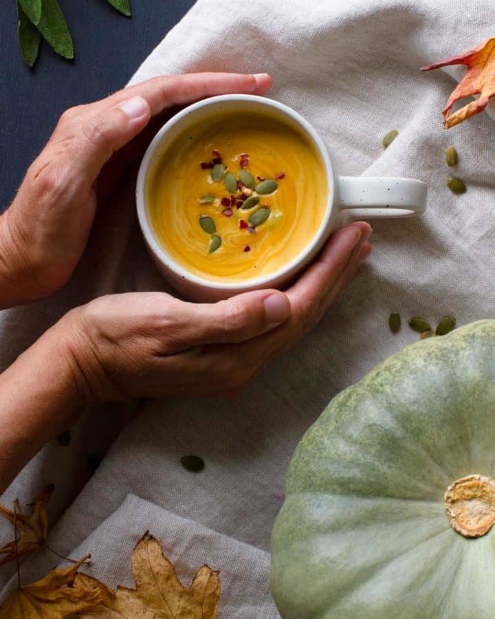 Two hands holding a white cup of pumpkin soup, cosy scene.