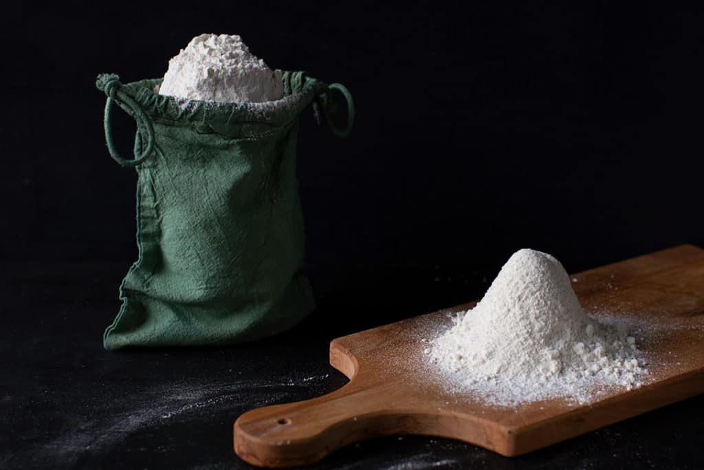 Flour shaped into a cone on a wooden board. Green linen bag with flour in the background. Moody scene.