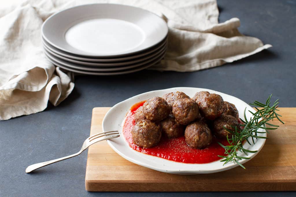 Meatballs without eggs, on tomato sauce, served on a white plate. A stack of plates and linen tea towel are in the background