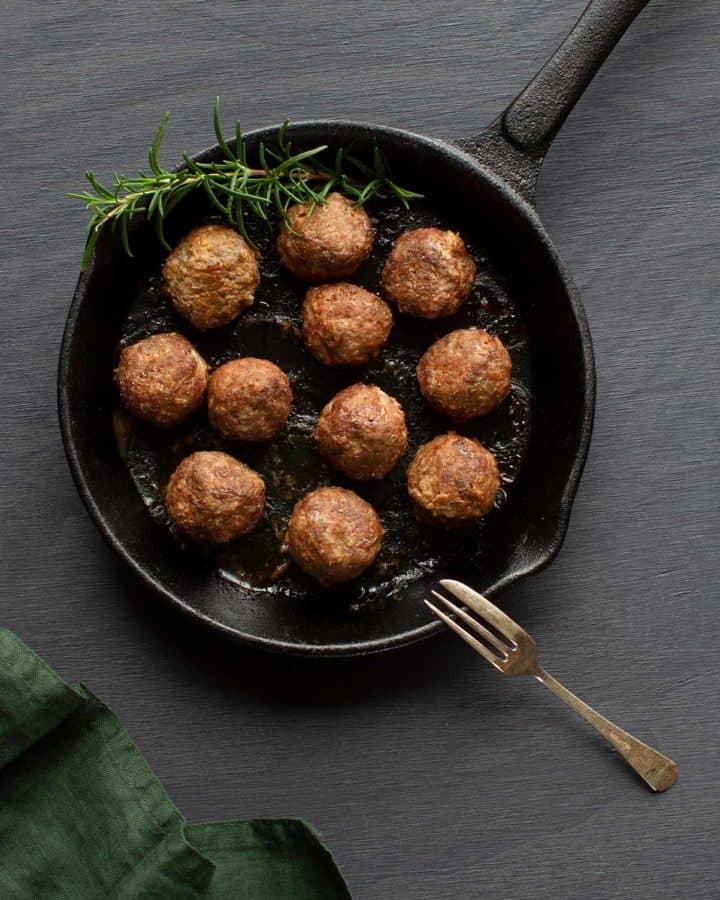 A flatlay of meatballs without eggs alongside a sprig of rosemary in a cast iron pan. A green tea towel is to the side. Moody scene.