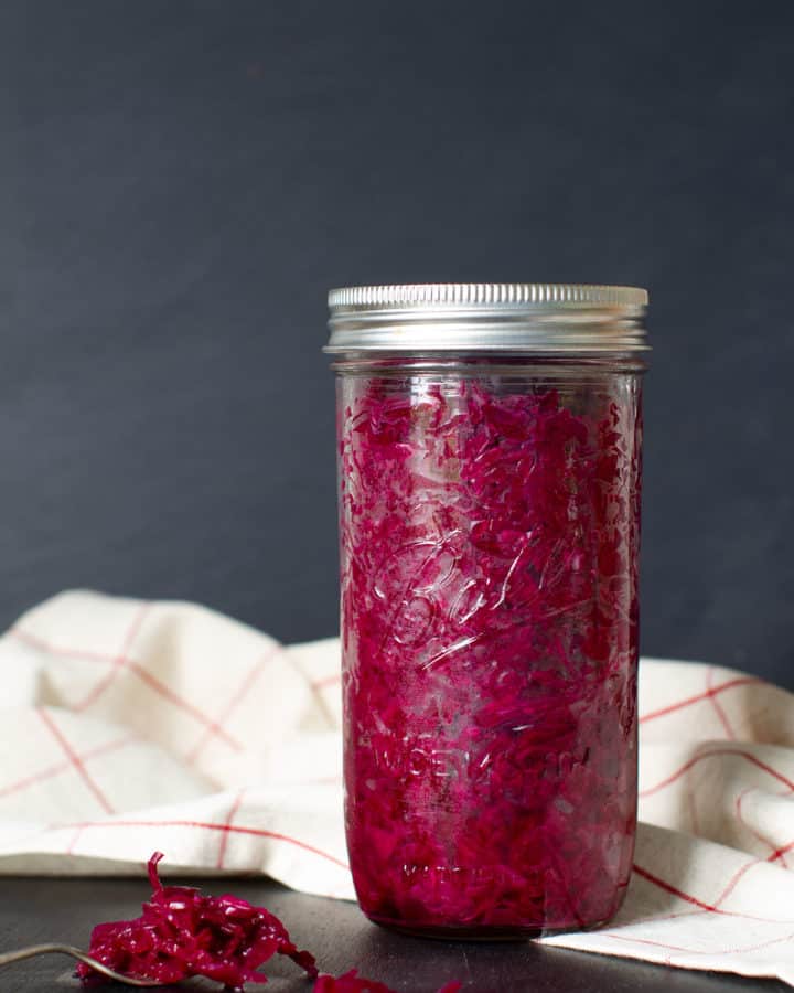 A jar of Red Sauerkraut Cabbage on a linen tea towel. A fork of sauerkraut in the foreground. Moody scene.