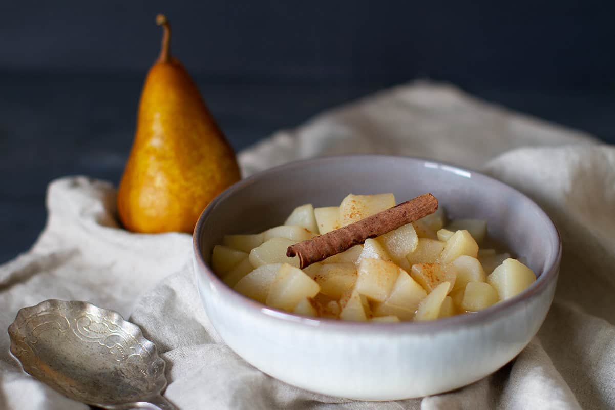 Close-up of a white bowl with stewed apples and pears with a cinnamon stick and a pear blurred in the background.