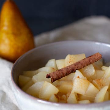 Close-up of a white bowl with stewed apples and pears with a cinnamon stick and a pear blurred in the background.
