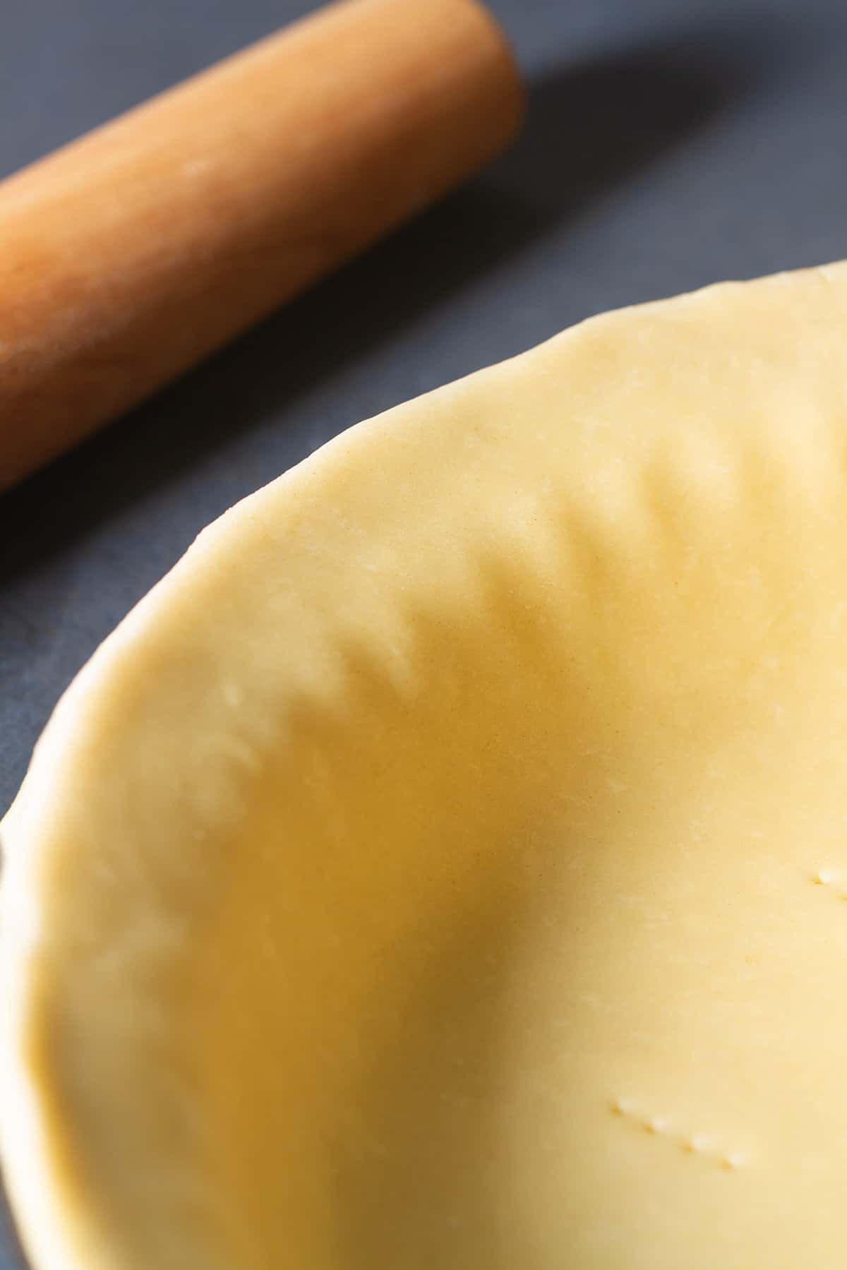 Close-up of a 3 ingredient pie crust in a pie tin next to a rolling pin on a dark background.