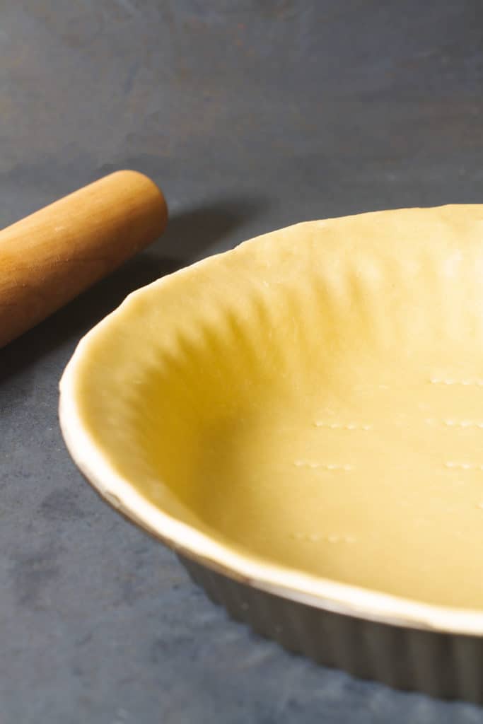 A 3 ingredient pie crust in a pie tin next to a rolling pin on a dark background.