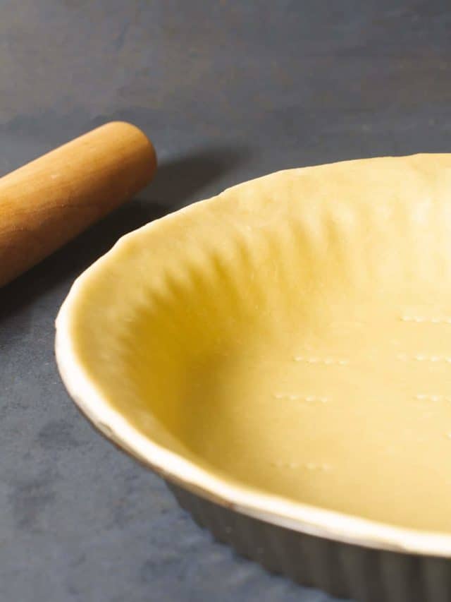 A 3 ingredient pie crust in a pie tin next to a rolling pin on a dark background.