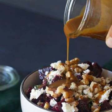 A bowl of roasted beetroot walnut and feta salad on a green tea towel. A jar of dressing is being poured onto the salad.