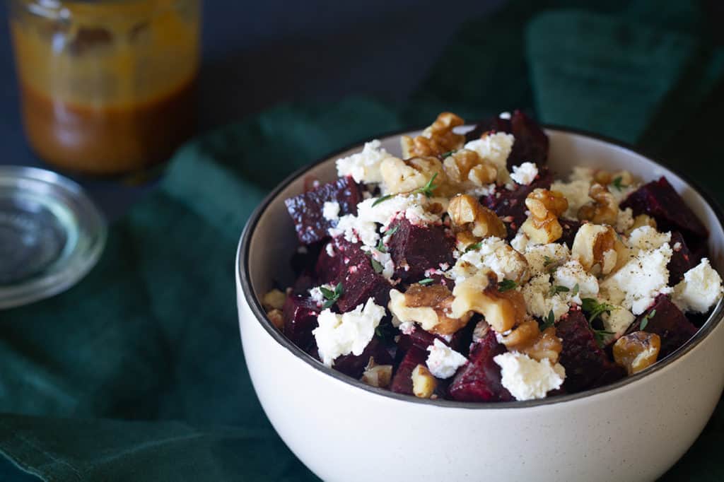 A bowl of roasted beetroot walnut and feta salad on a green tea towel. A jar of dressing is blurred out in the background.