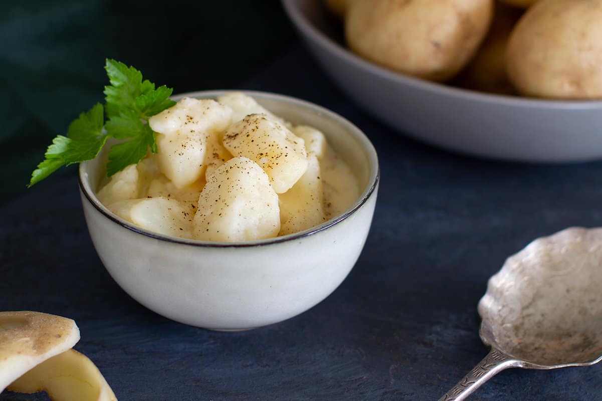 A small bowl of stewed potatoes with parsley. A bowl of potatoes is in the background.