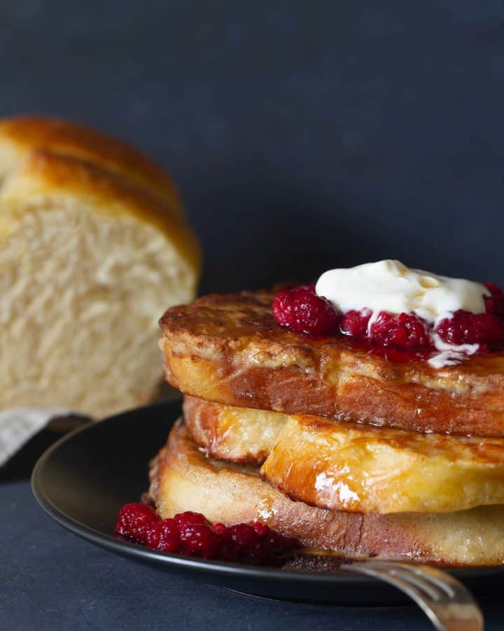 A stack of sourdough french toast on a black plate, topped with raspberries and yogurt. A brioche loaf is in the background.
