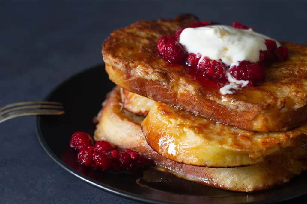 A stack of sourdough french toast on a black plate, topped with raspberries and yogurt.