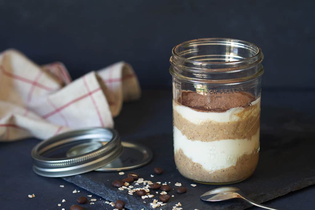 A clear jar of tiramisu overnight oats on a shingle plate. Mason jar lid, tea towel, spoon, oats, and coffee beans are next to it.