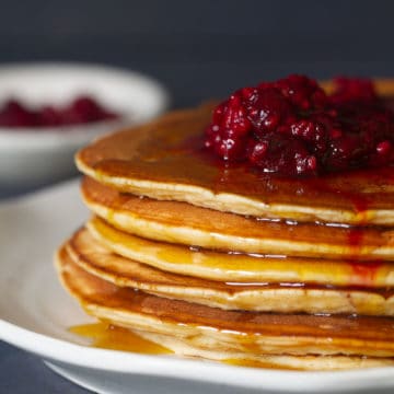 A stack of whey pancakes with raspberries and maple syrupon a white plate next to a small bowl of raspberries and a tea towel.