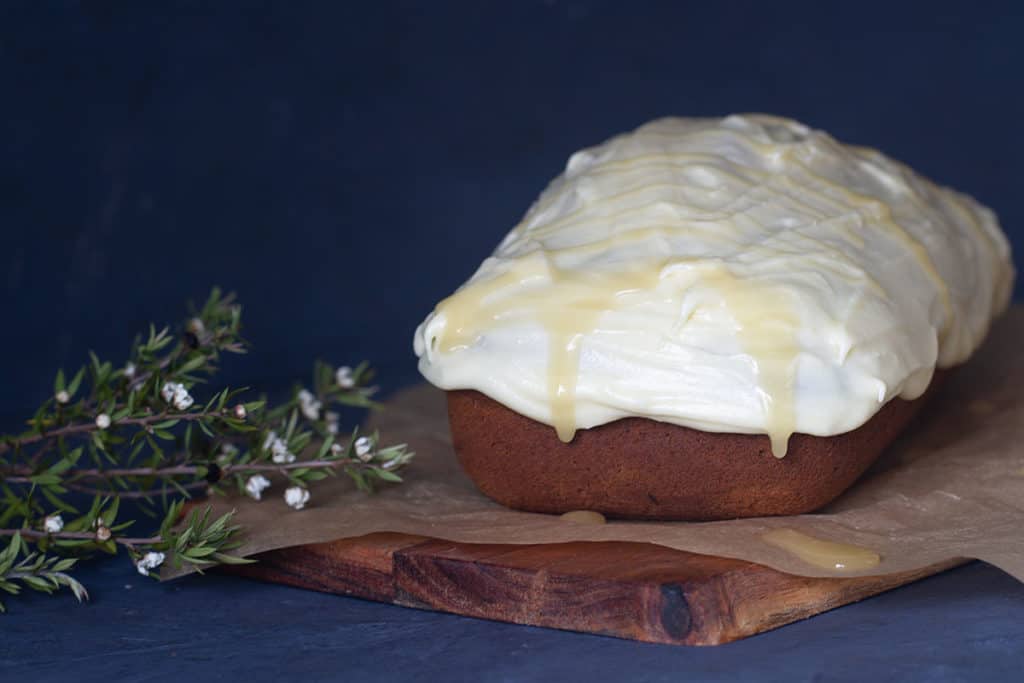 A spiced honey loaf cake on a wooden board next to a Manuka tree sprig.