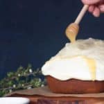 A spiced honey loaf cake on a wooden board. Honey drizzle is added with a honey spoon.