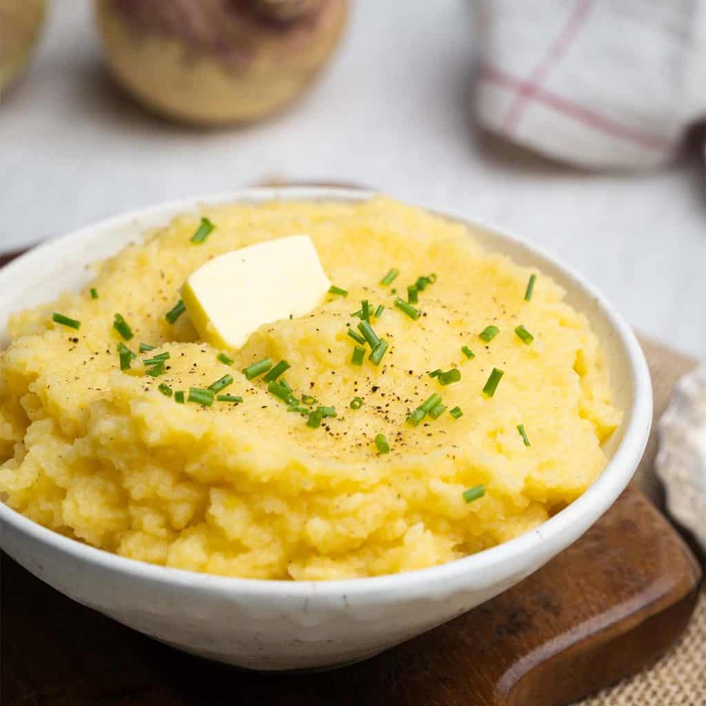 A bowl of extra creamy swede mash on a wooden board, next to swedes and a tea towel.