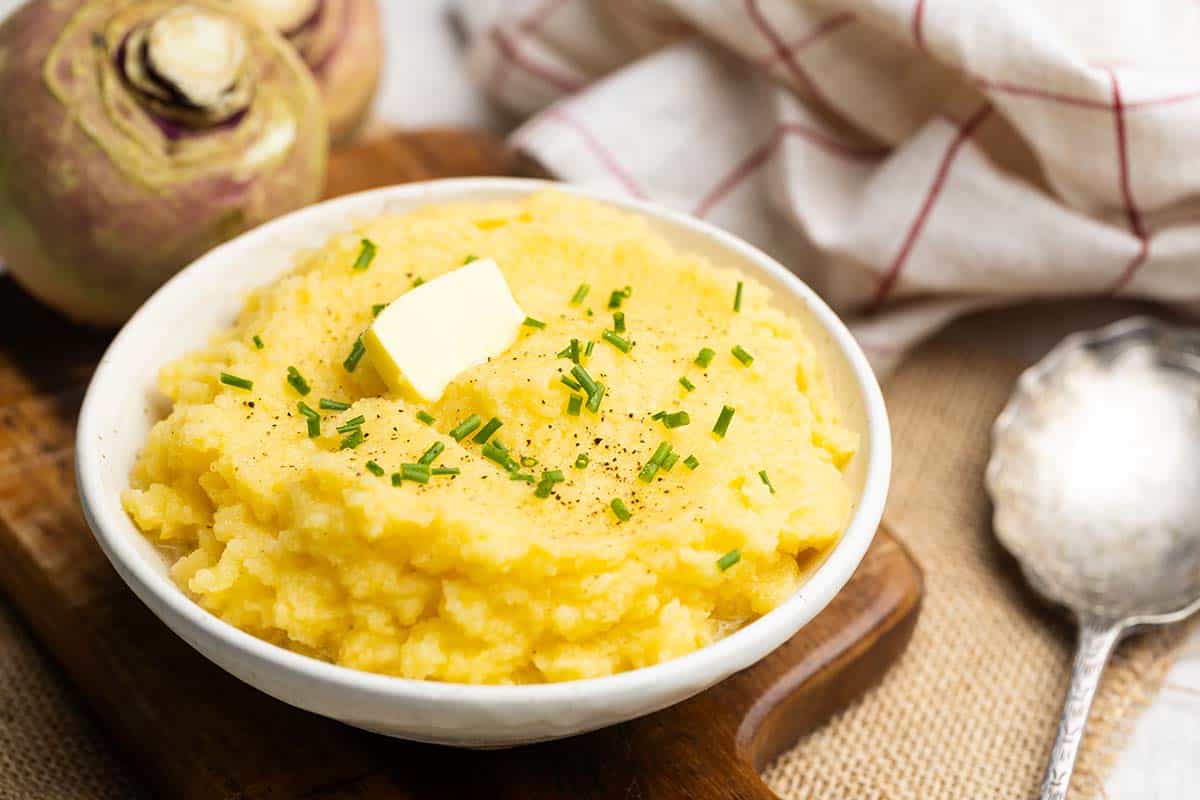 A bowl of extra creamy swede mash on a wooden board, next to swedes and a tea towel.