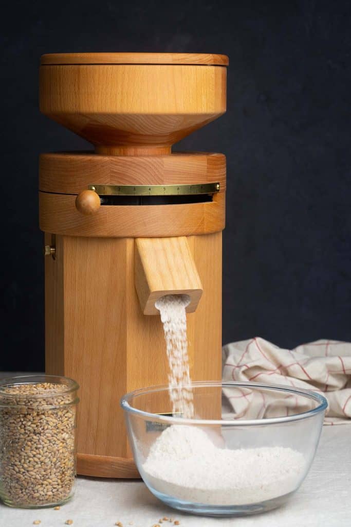 A jar of grains next to a bowl with flour and a grain mill in the background milling flour.