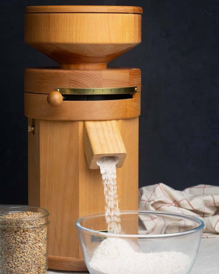 A jar of grains next to a bowl with flour and a grain mill in the background milling flour.