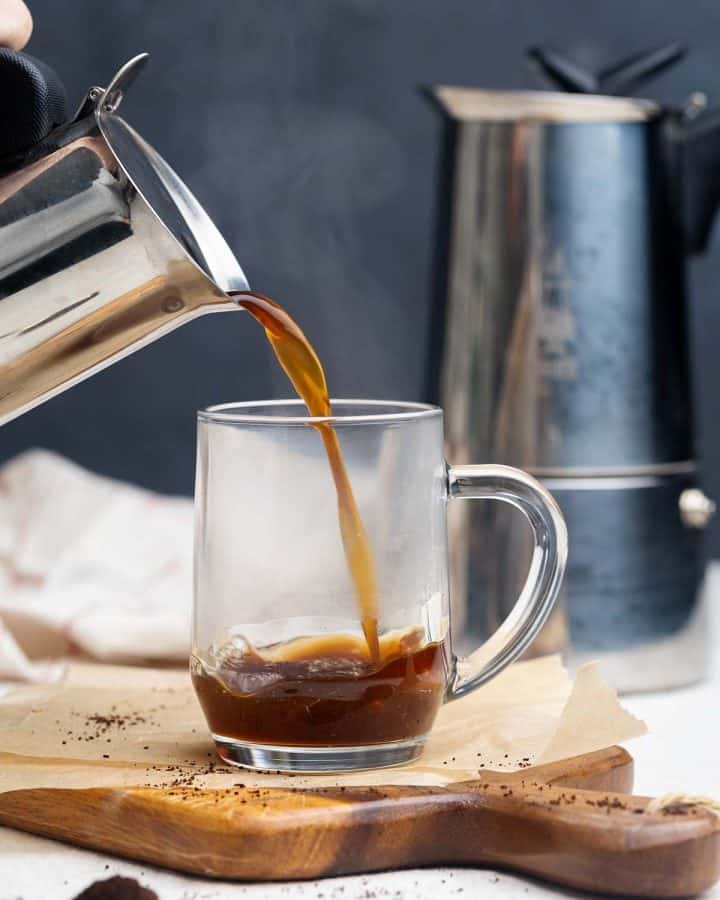 A Moka pot pouring coffee into a glass cup.