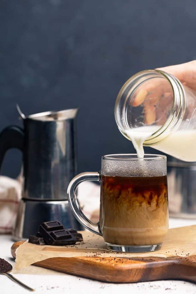 Milk being poured into a glass cup of coffee, Moka pot in the background.