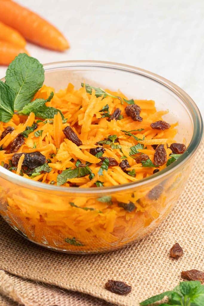 A glass bowl filled with raw carrot salad, garnished with mint and raisins.