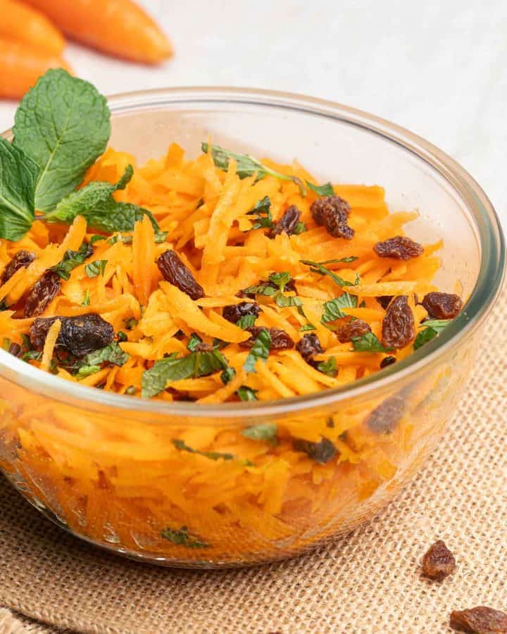 A glass bowl filled with raw carrot salad, garnished with mint and raisins.