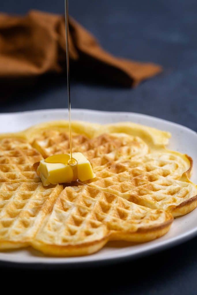A fluffy American waffles on a plate being drizzled with maple syrup.