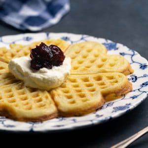 A thin and crispy Swedish waffle on a patterned plate, topped with cream and jam. Moody scene.
