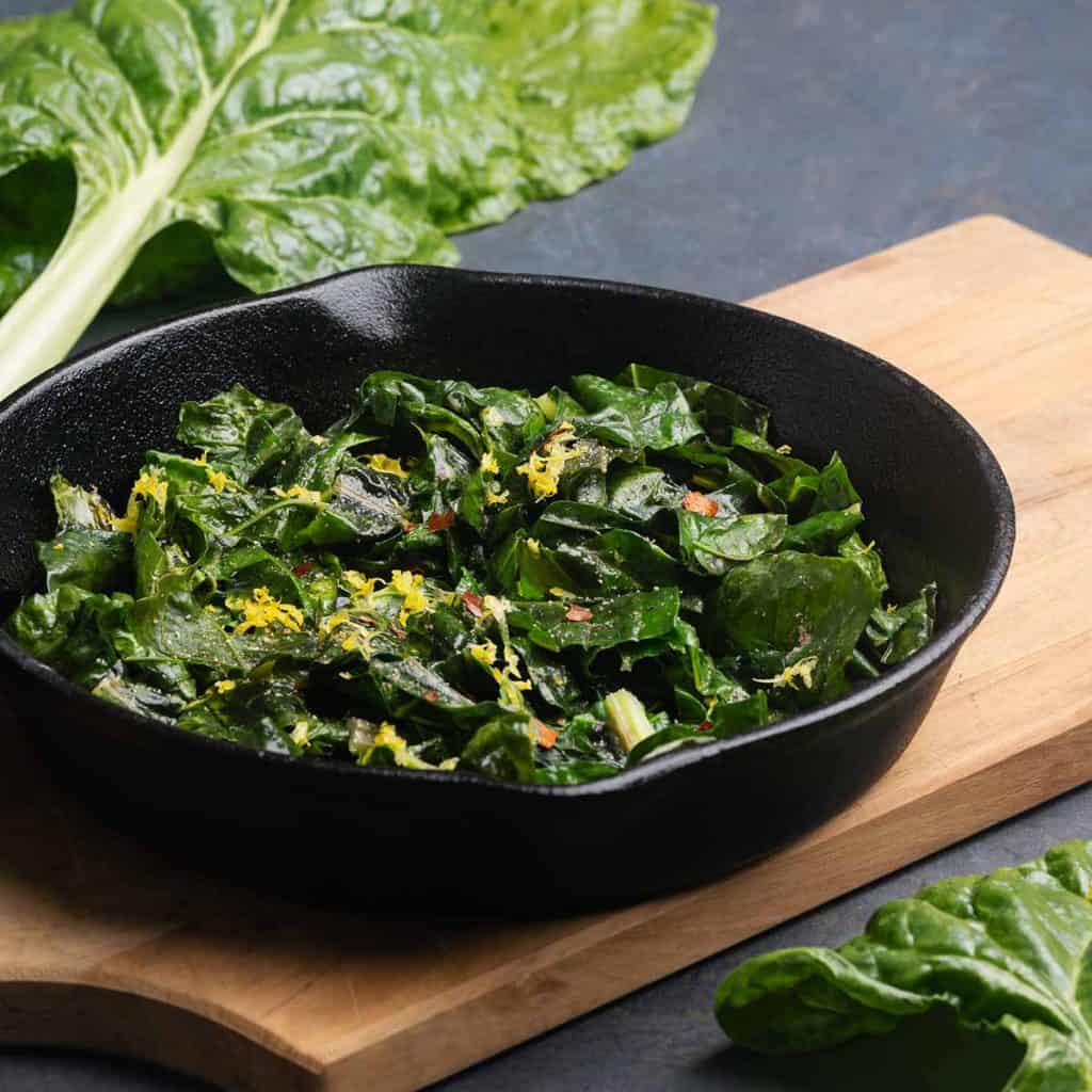 Sautéed silverbeet in a cast iron skillet on a wooden board next to silverbeet stems.