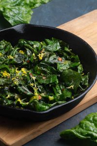 Sautéed silverbeet in a cast iron skillet on a wooden board next to silverbeet stems.