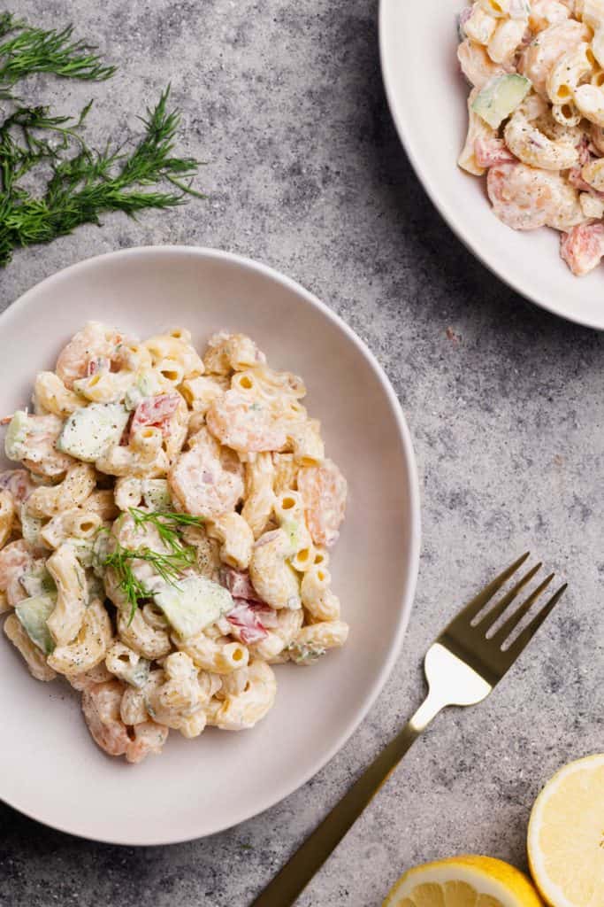 A white bowl filled with shrimp macaroni salad next to lemon halves and dill.