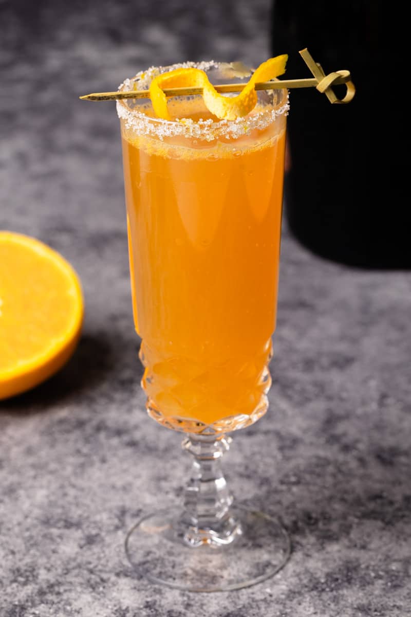 A glass of Rosé mimosa on a slate next to an orange and a bottle of Rosé champagne.