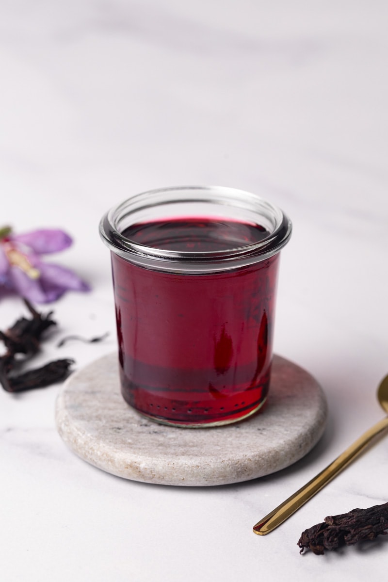 Homemade hibiscus syrup in a glass jar on a marble coaster.