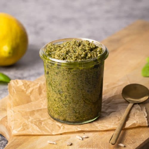 Basil sunflower seed pesto in small glass jar on wooden board next to sunflower seeds, a golden spoon, basil, and a lemon.