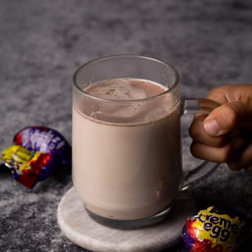 Cadbury creme egg hot chocolate in a glass cup next to creme eggs.