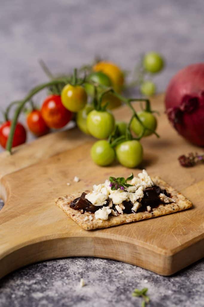 A cracker topped with caramelized onion tomato jam and feta on a wooden board next to a jar of jam.