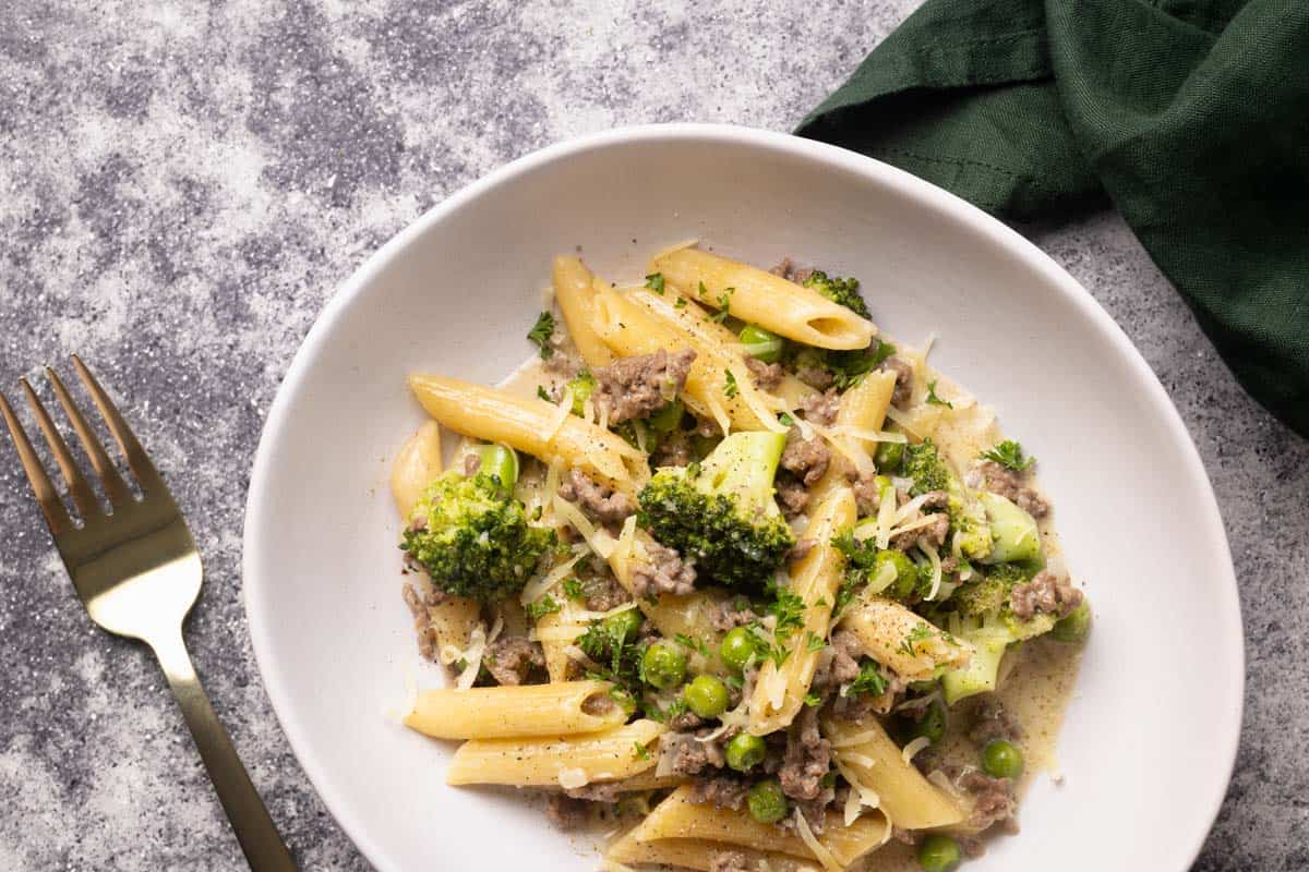 A white bowl filled with ground beef Alfredo pasta next to a large pan.