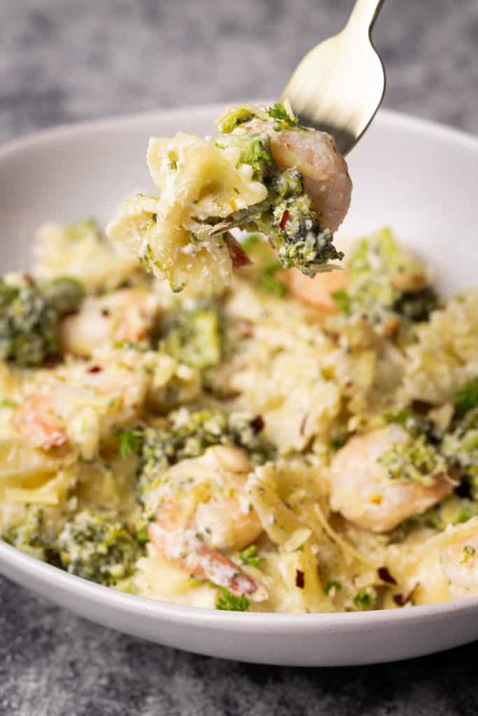 A fork with a bowtie pasta, a shrimp, and broccoli floret.