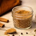 A jar of homemade Masala chai spice mix next to whole ingredients and a small spoon.