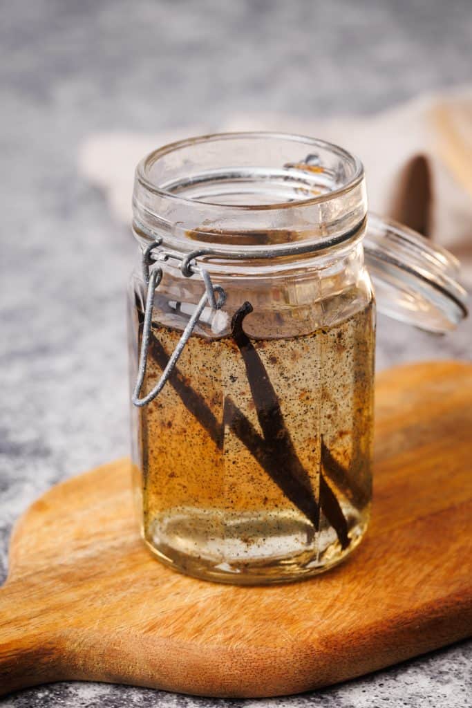 Vanilla syrup in a glass container on a wooden board next to a golden spoon and a linen napkin.