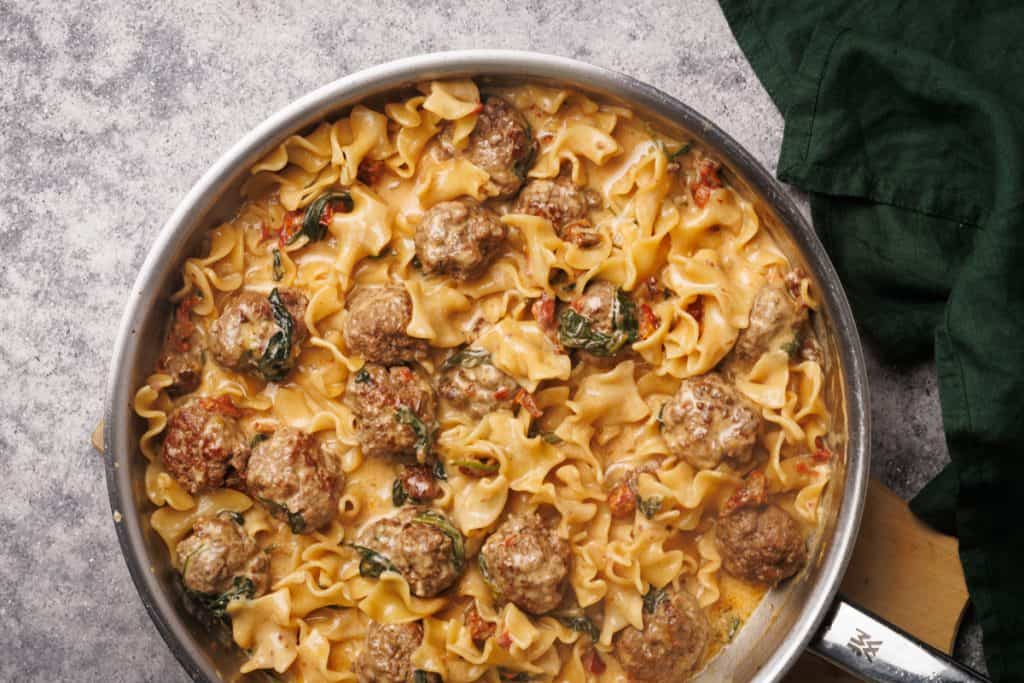 A skillet filled with creamy pasta, meatballs, and spinach, garnished with a rich sauce. The dish is set on a textured gray surface next to a dark green cloth.