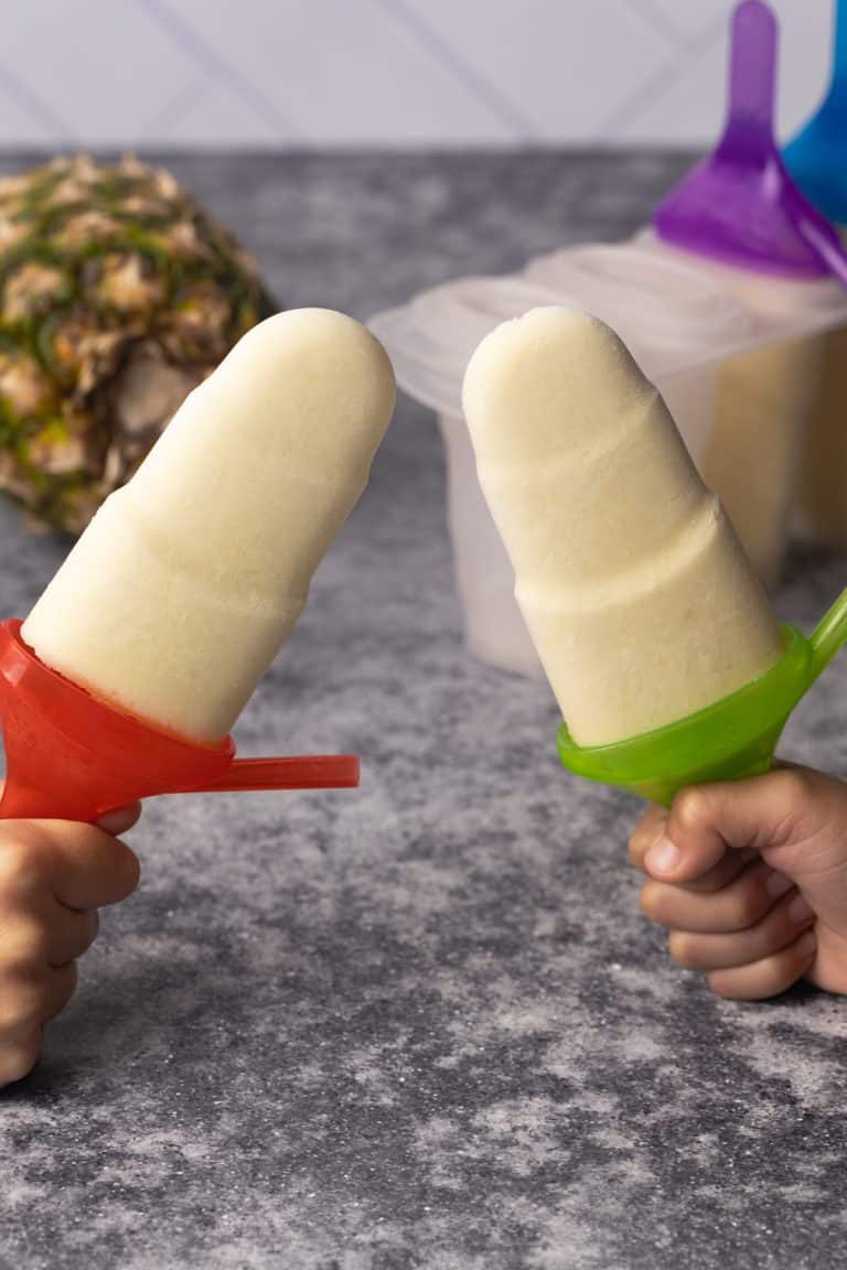 Tropical popsicles held by kids' hands next to a pineapple and ice block moulds.