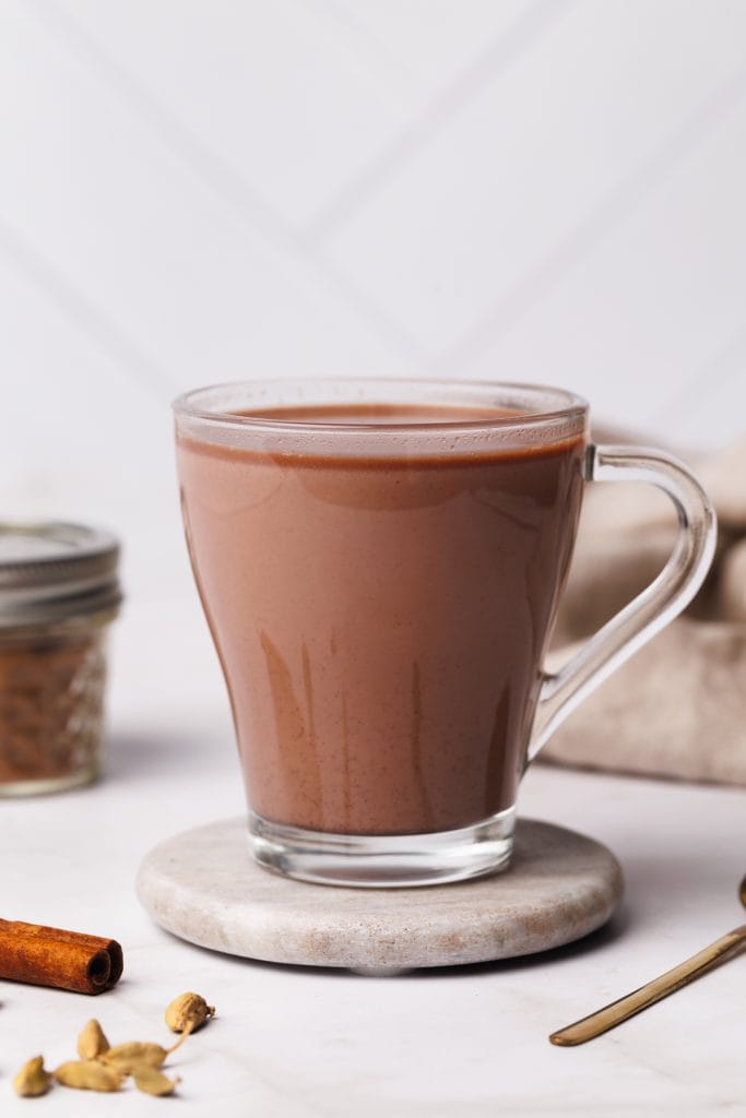 A glass cup filled with chai hot chocolate on a marble coaster next to a jar of spice mix and a linen tea towel.