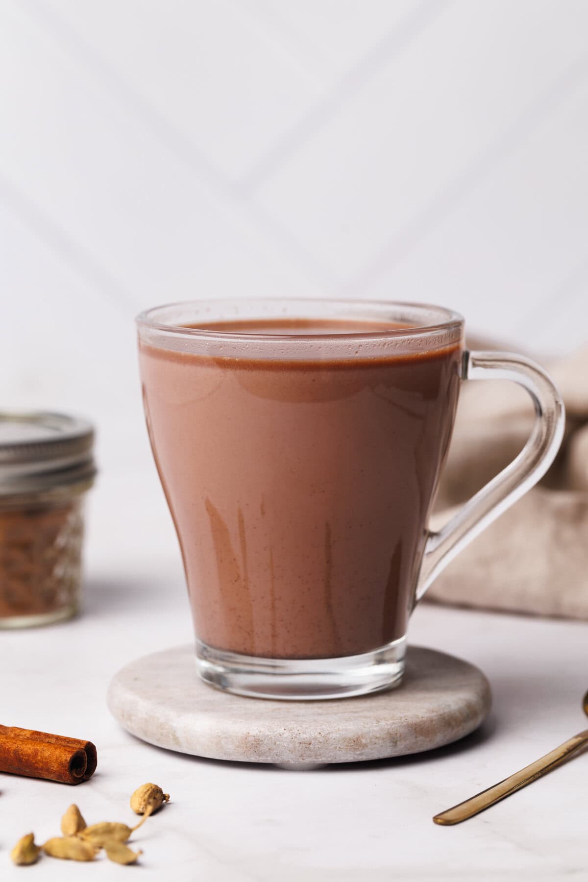 A glass cup filled with chai hot chocolate on a marble coaster next to a jar of spice mix and a linen tea towel.