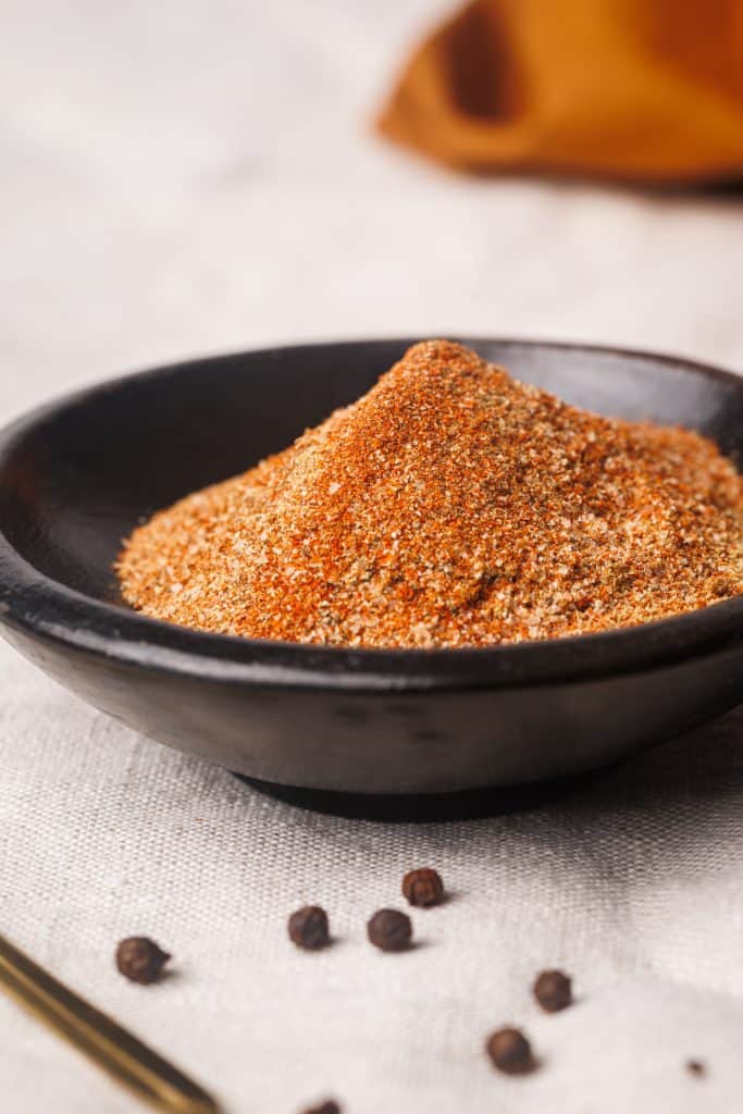 Lamb dry rub in a black bowl next to pepper kernels.