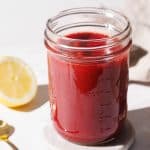 A mason jar filled with strawberry puree on a marble coaster next to a golden spoon and half a lemon.