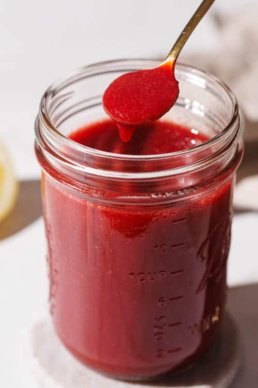 A mason jar filled with strawberry puree on a marble coaster next to a golden spoon and half a lemon.