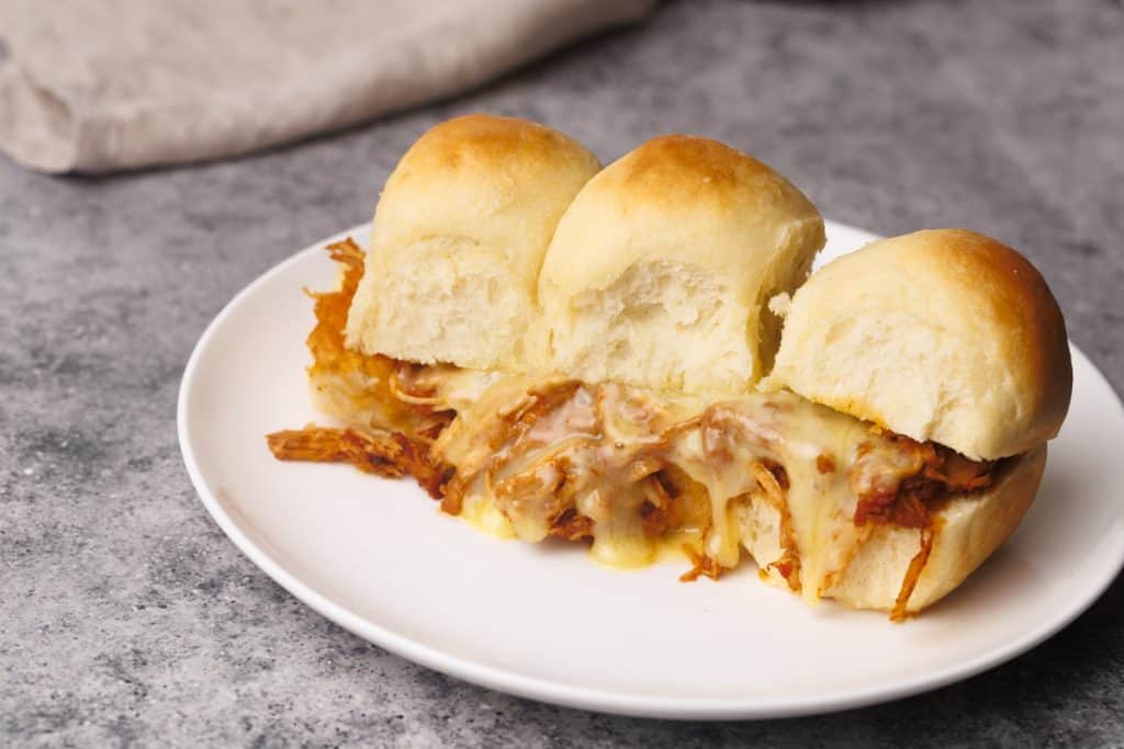 Three pull-apart rolls filled with shredded, cheesy meat are served on a white plate atop a gray textured surface. A beige cloth is partially visible in the background.