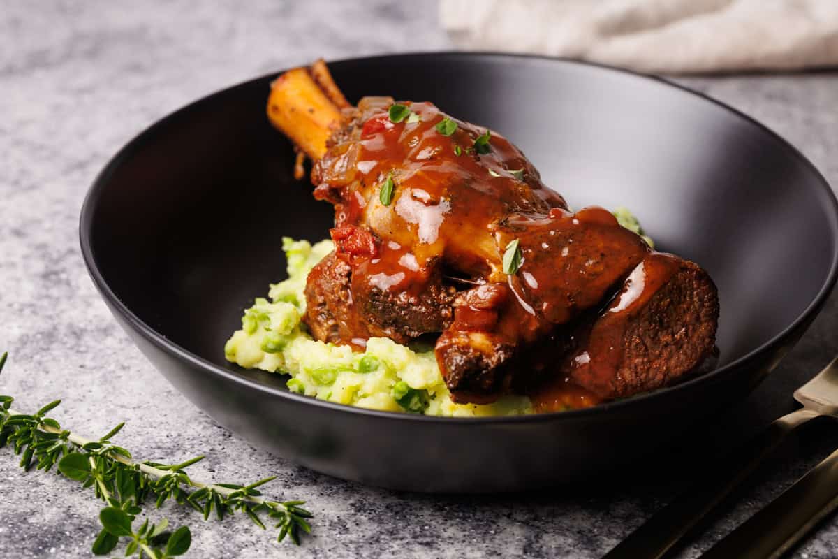 Lamb shanks on mashed peas and potatoes in a black bowl.